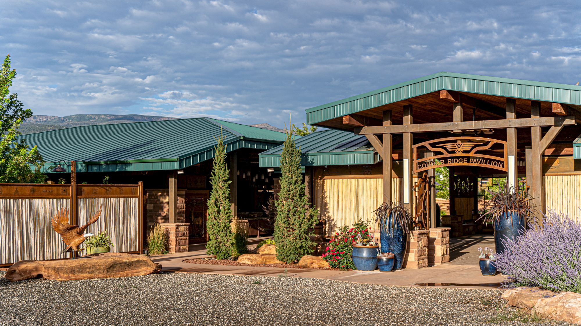 A rustic building with a green roof, wooden accents, and surrounding plants and gravel under a cloudy sky.