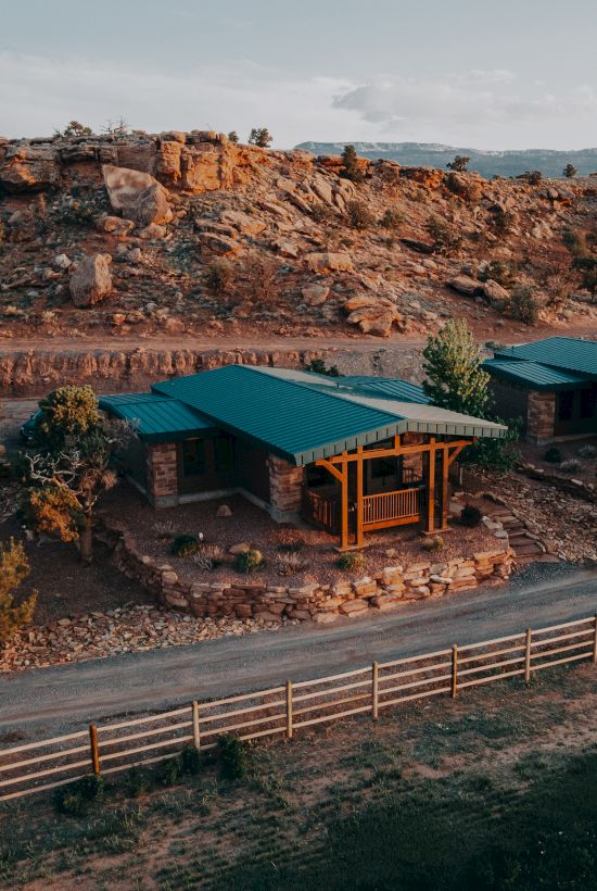 A cozy cabin with a green roof is nestled among rocky hills and desert vegetation, with a wooden fence lining a dirt road in the foreground.