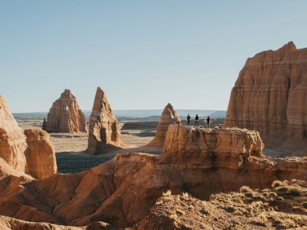 A desert landscape with rock formations and three people standing on a ledge in the middle of the scene, under a clear blue sky.