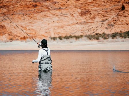 A person is standing in water wearing fishing gear and casting a line, with rocky terrain in the background.