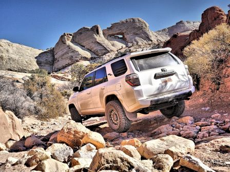 A white SUV navigates a rocky, off-road terrain with rugged hills in the background and a clear blue sky.