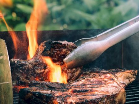 Grilling steaks over an open flame with tongs, surrounded by greenery in the background, creating a lively outdoor cooking scene.