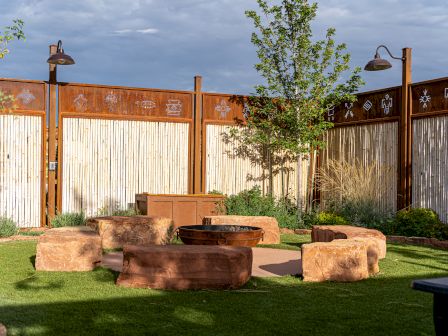 Outdoor space with grass, stone seating around a fire pit, wooden fence with artistic details, and overhead lighting.