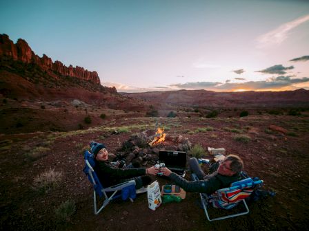 Two people sit in chairs by a campfire in a desert landscape at sunset, enjoying a scenic view.