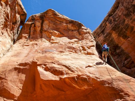 A person is rock climbing on a steep, reddish-brown rock face under a clear blue sky, using climbing gear and a rope for safety.