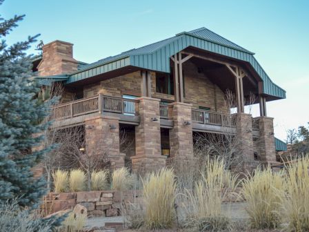 A large house with stone walls, wooden accents, and a green metal roof, surrounded by tall grasses and trees.