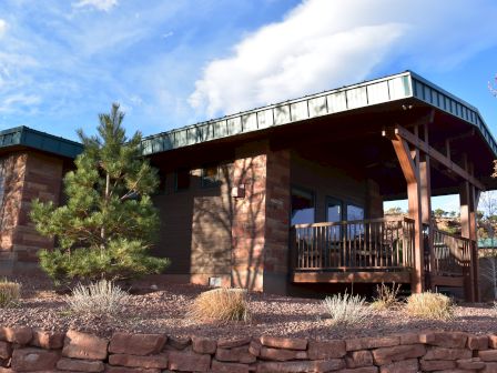 A wooden house with a porch, surrounded by trees and bushes, under a partly cloudy sky.