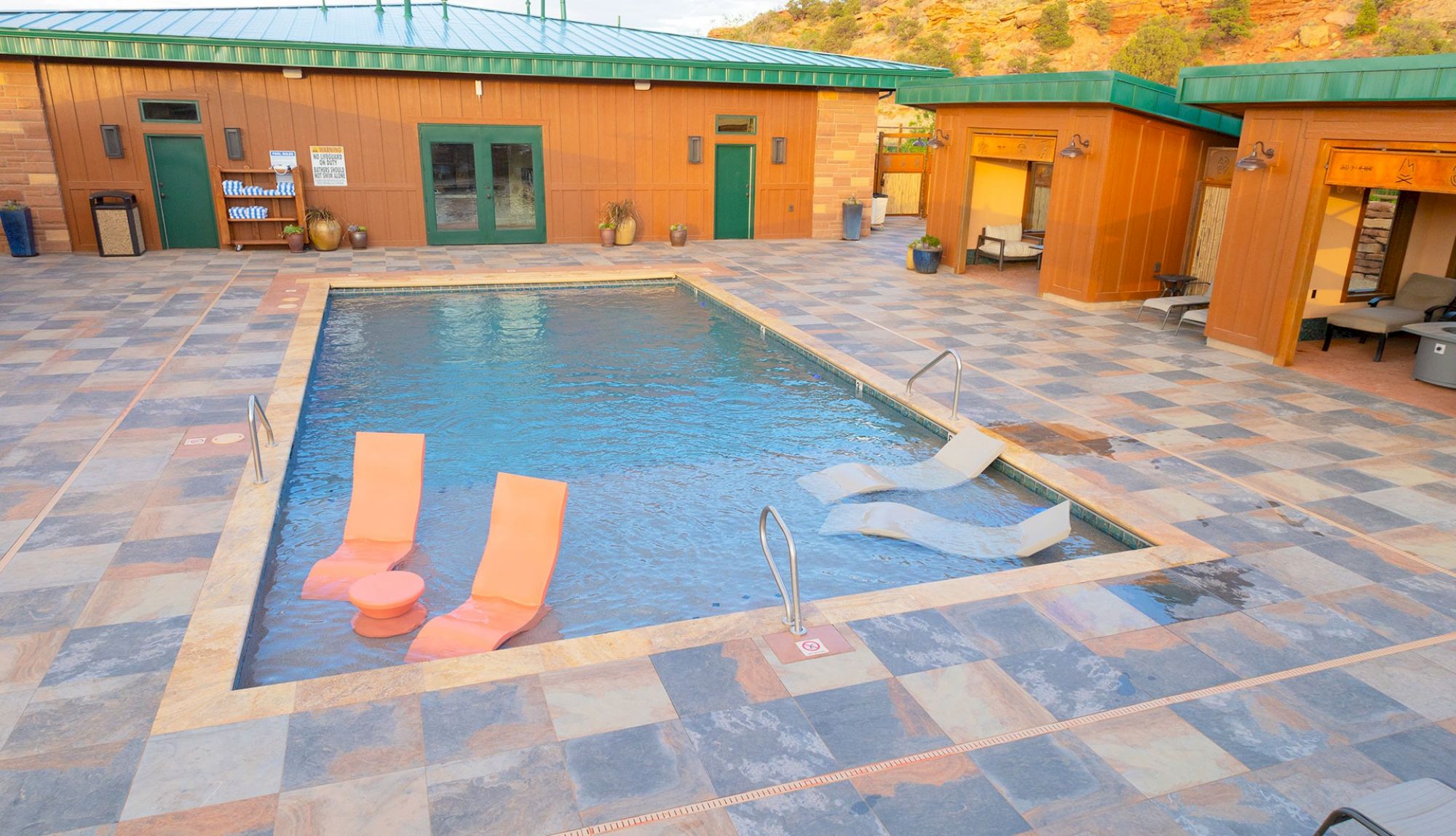 A poolside area with lounge chairs in the water, surrounded by modern buildings and a tiled deck.