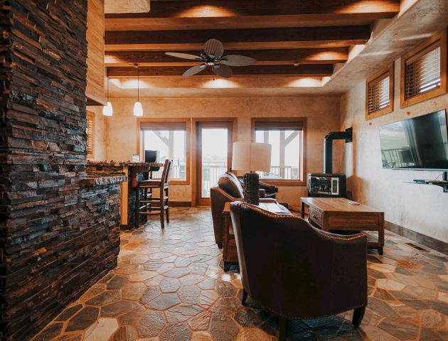 A cozy living room featuring stone accents, a ceiling fan, wooden beams, a TV, and comfortable seating near large windows.