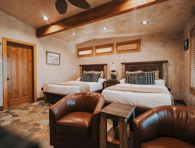The image shows a cozy bedroom with two beds, leather chairs, rustic decor, and wooden elements, creating a warm and inviting atmosphere.