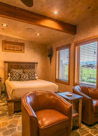 A cozy room with a bed, two leather chairs, a table, plants, and a window view, featuring warm lighting and a rustic design.