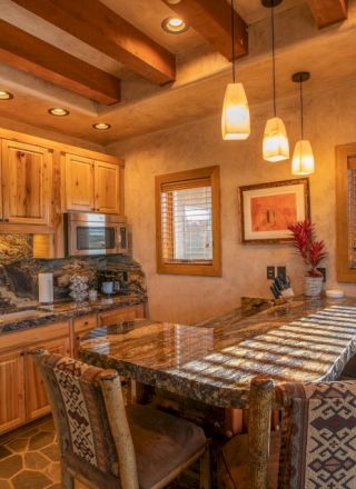 A rustic kitchen with wood cabinets, stone accents, hanging lights, and a counter with patterned chairs is shown.