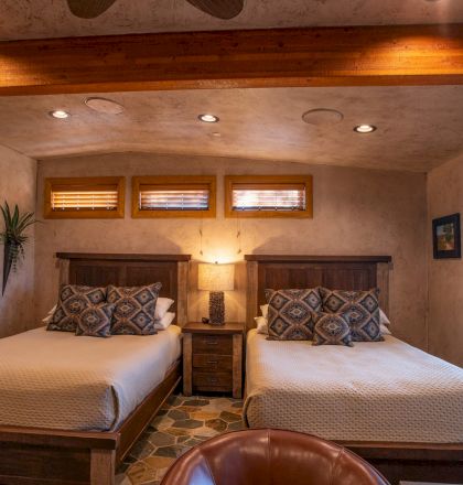 A cozy bedroom with two beds, patterned pillows, a nightstand with a lamp, and warm lighting. There are artworks and a window.