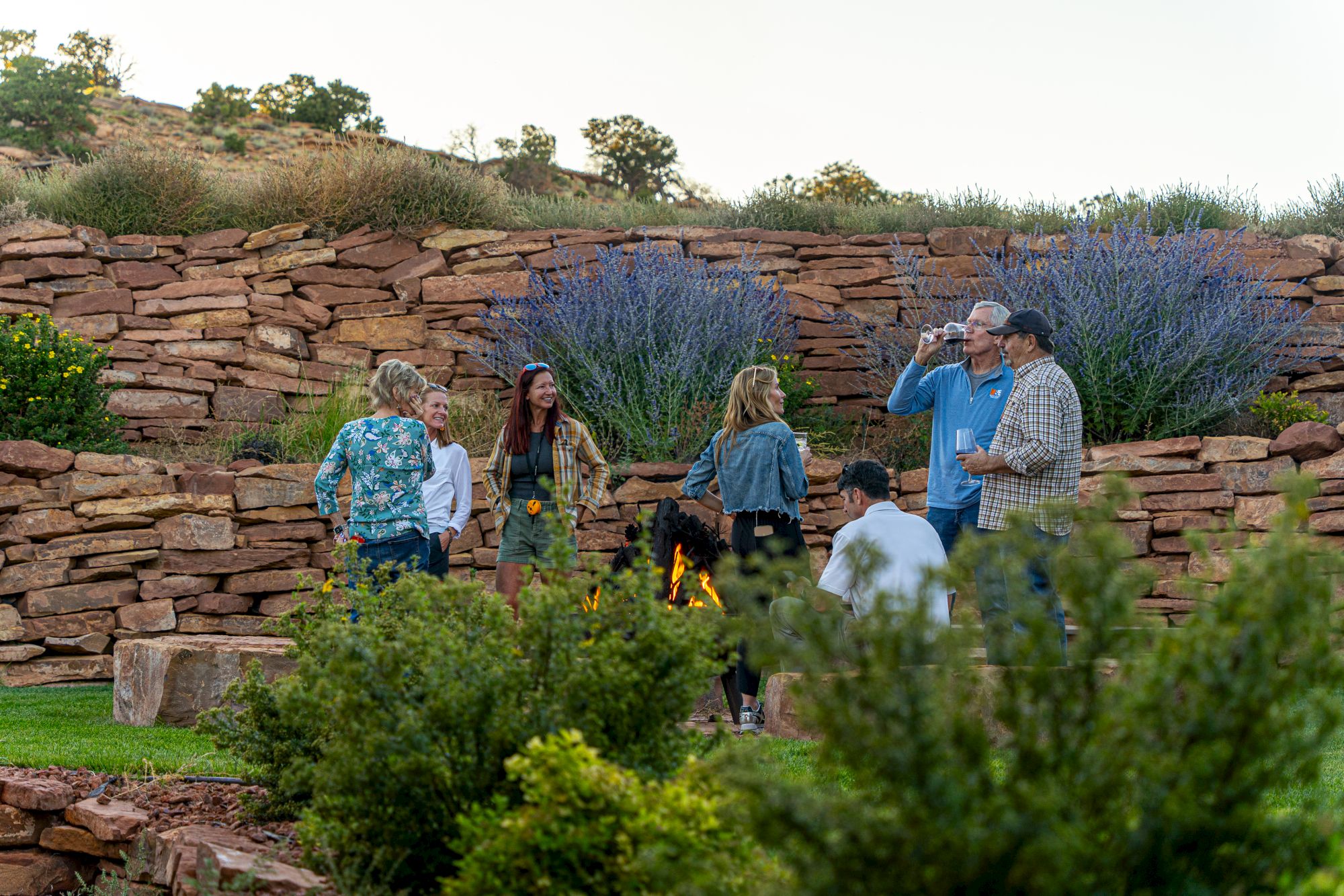 A group of people are standing and talking in a landscaped garden with stone walls and greenery around them.