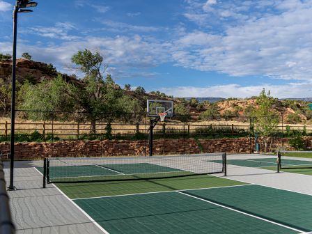 An outdoor sports area featuring a pickleball court and basketball hoop, set against a scenic backdrop of trees and rocky hills under a blue sky.