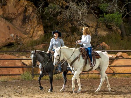 Two people are riding horses in an outdoor setting, surrounded by rocks and trees, with a metal fence in the background.
