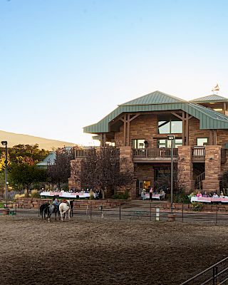 A ranch scene with a large brick building, horses in a fenced area, and dining tables set up outdoors under a clear sky.