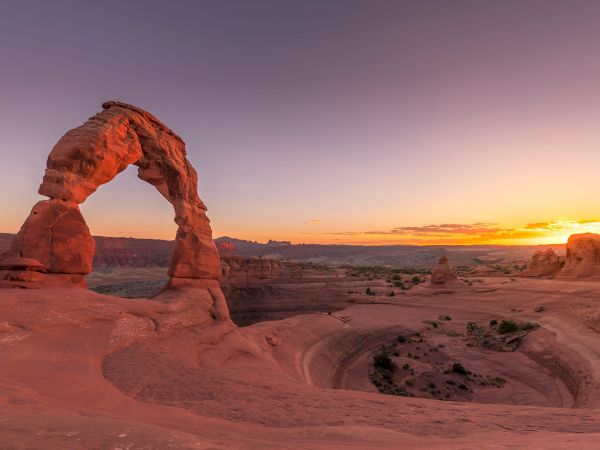 The image shows Delicate Arch in Arches National Park at sunset, with a clear sky and vast desert landscape surrounding it.