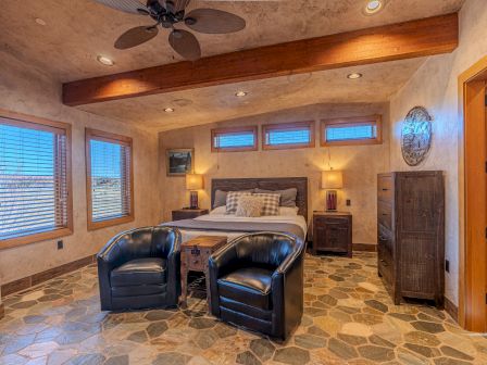 A cozy bedroom with a bed, two chairs, a ceiling fan, and warm lighting, featuring wooden accents and large windows.