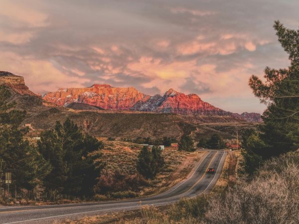 A winding road leads to vibrant, sunlit mountains beneath a colorful sky, flanked by trees and open landscape.
