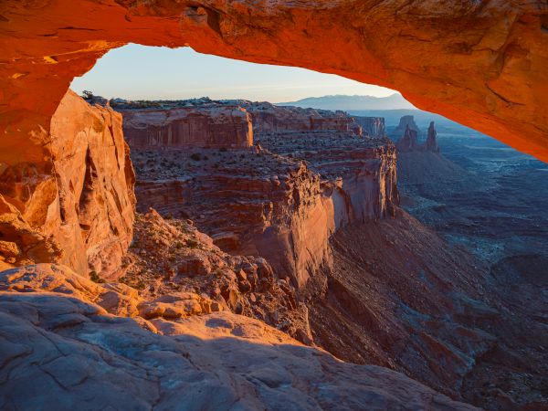 A scenic view of a rock arch with canyon cliffs and a valley in the background, illuminated by sunlight in a desert landscape.