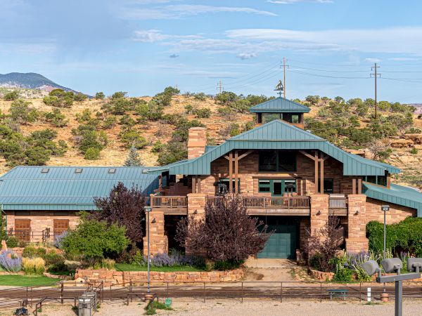 A large, rustic building with a green roof set in a desert landscape with shrubs and hills in the background.