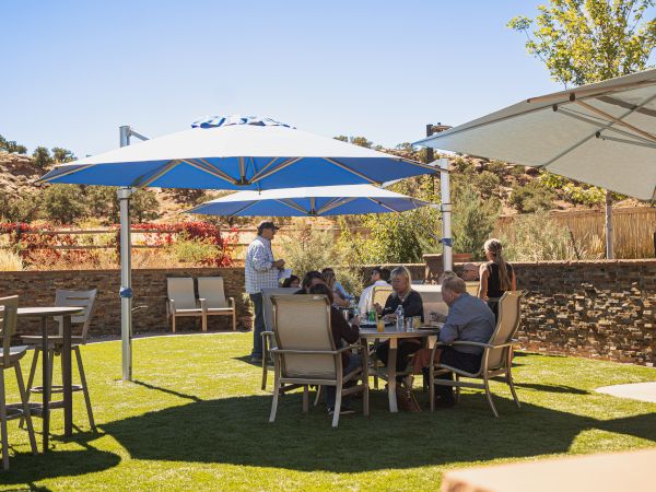 People are sitting and standing in a sunny outdoor area with tables, chairs, and umbrellas on a lawn, surrounded by a scenic landscape.