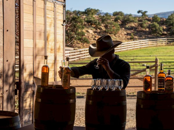 A person in a cowboy hat stands at barrels with bottles and glasses, set against a rustic outdoor backdrop.