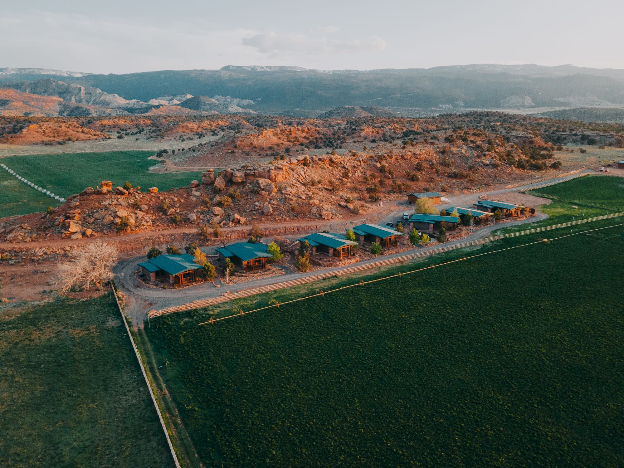 An aerial view of a rural area with houses lined up near rocky hills and expansive green fields in the foreground.