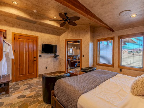 A cozy bedroom with a large bed, wall-mounted TV, wooden accents, ceiling fan, and windows letting in natural light.