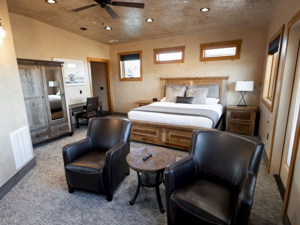 The image shows a cozy bedroom with a large bed, two armchairs, a wooden wardrobe, side tables, and a ceiling fan in a warmly lit space.