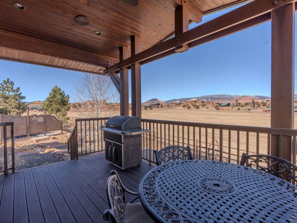 A covered patio with a metal dining table and a grill overlooks a vast, open landscape with trees and hills under a clear blue sky.