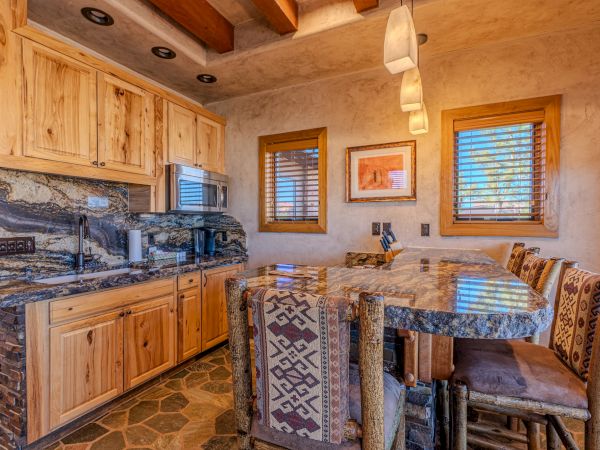 A rustic kitchen with wooden cabinets, a stone countertop, and patterned chairs. Two windows and pendant lights add warmth to the space.