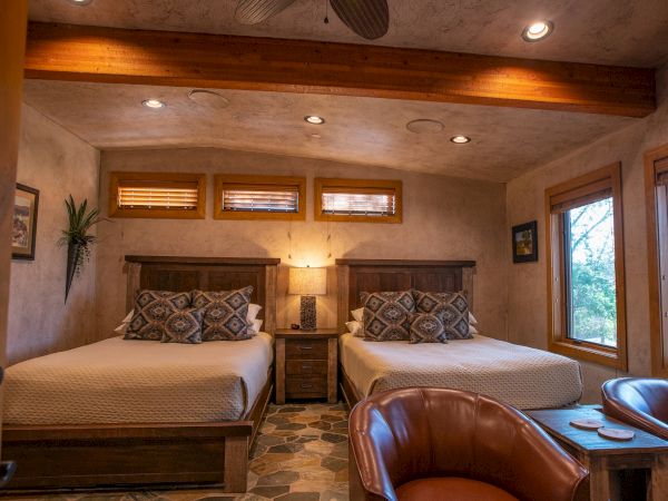 A cozy bedroom features two beds, warm lighting, wooden beams, and a leather chair, creating a welcoming atmosphere.
