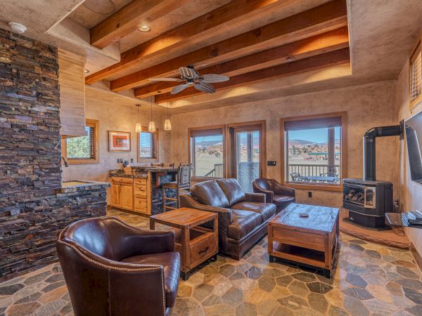 A rustic living room with leather seating, wooden beams, stone accents, a TV, and large windows with scenic views.