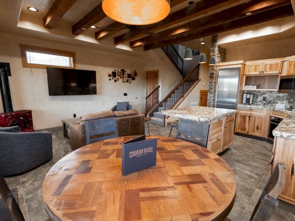 A modern living space features a wooden dining table, cozy couches, wall-mounted TV, kitchen area, and visible staircase.