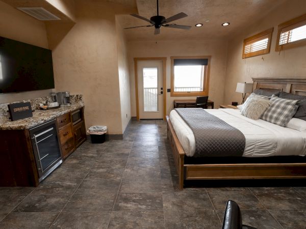 A cozy bedroom with a large bed, wooden furniture, a TV, and a kitchenette area, featuring neutral tones and modern decor.