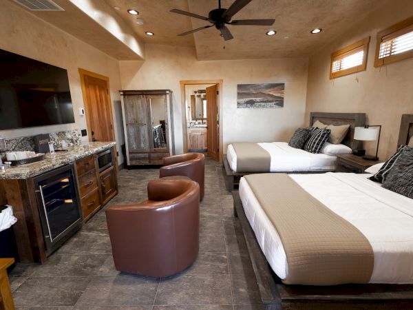 The image shows a hotel room with two beds, a large TV, a kitchenette, brown chairs, and a modern rustic design.