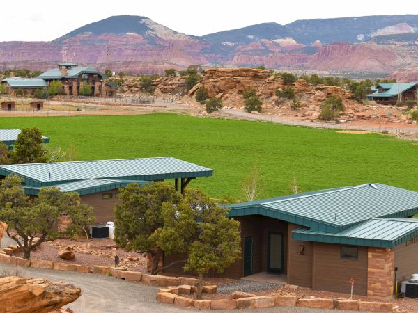 The image shows cabins with green metal roofs surrounded by trees and a large grassy area, set against a backdrop of mountains and red rocks.