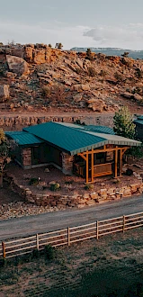 A small house with a green roof is nestled in a rocky, desert landscape, surrounded by a wooden fence and sparse vegetation.